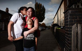 A woman embraces two children outside a row of terraced houses on a residential street.