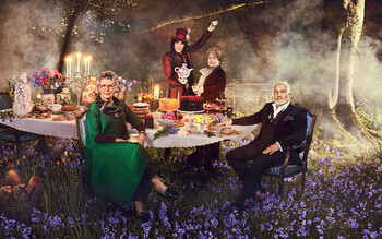 Judges Paul Hollywood and Prue Leith sit at a banquet table full of food for a Mad Hatter's Tea Party in a woodland grove. Hosts Noel Fielding and Sandi Toksvig stand behind with a teapot.