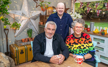 Judges Paul Hollywood and Prue Leith sit with mugs at a table inside the Bake Off Tent. Host Matt Lucas stands behind them near a large star decoration.