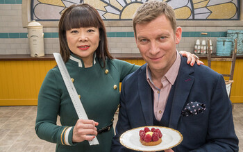 Judge Cherish Finden stands with a ruler on set with her arm around fellow judge Benoit Blin, who holds a patisserie-style tart.