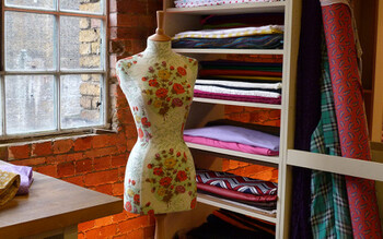 A dressmaking mannequin stands in the sewing room near a window and shelves of fabric.