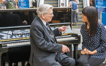 Claudia Winkleman sits with a piano player at a piano in St Pancras station. 